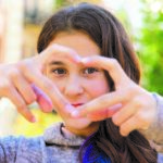 Mädchen mit langen braunen Haaren und braunen Augen formt mit seinen Fingern ein Herz und schaut hindurch in die Kamera.