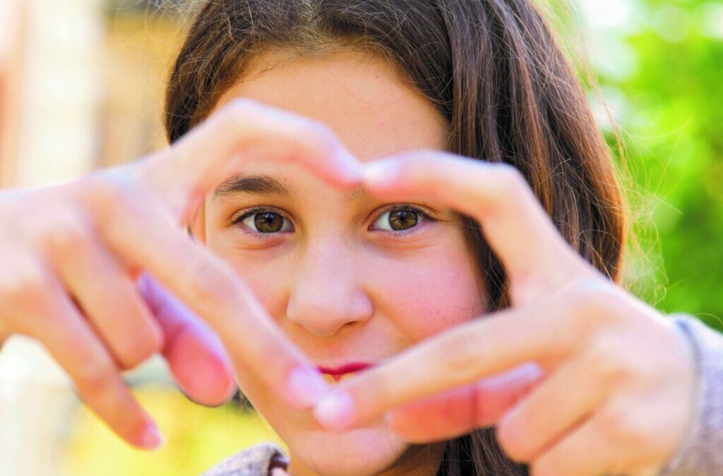 Mädchen mit langen braunen Haaren und braunen Augen formt mit seinen Fingern ein Herz und schaut hindurch in die Kamera.
