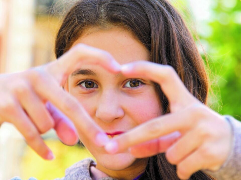 Mädchen mit langen braunen Haaren und braunen Augen formt mit seinen Fingern ein Herz und schaut hindurch in die Kamera.