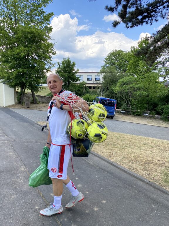 Fußballtrainer Toni mit Bällen bepackt auf dem Weg zum Fußballtraining mit den Kids