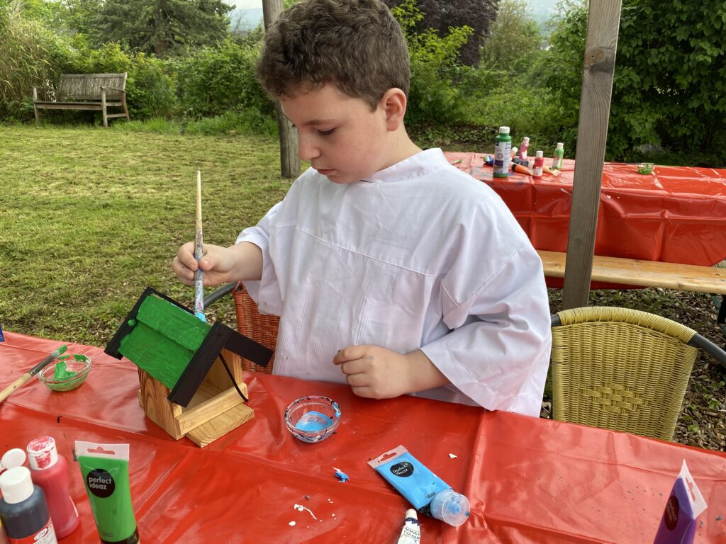 Junge in weißem Malerkittel malt Vogelhäuschen grün und schwarz an