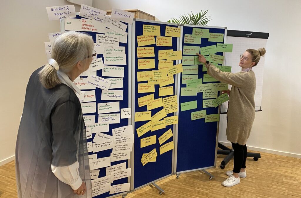 Referentin mit weißen Haaren und grauer Strickjacke und Veranstaltungsteilnehmerin mit blondem Dutt, Brille und beigem Strickkleid stehen vor einer Moderationswand mit vielen weißen, gelben und grünen Moderationskärtchen