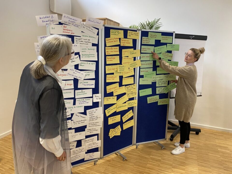 Referentin mit weißen Haaren und grauer Strickjacke und Veranstaltungsteilnehmerin mit blondem Dutt, Brille und beigem Strickkleid stehen vor einer Moderationswand mit vielen weißen, gelben und grünen Moderationskärtchen