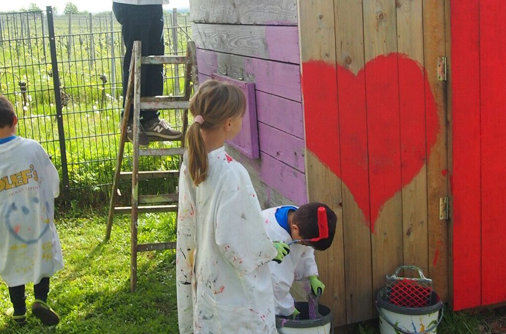 Blondes Mädchen in Malerkittel steht vor einer Holzhütte und malt ein großes rotes Herz darauf